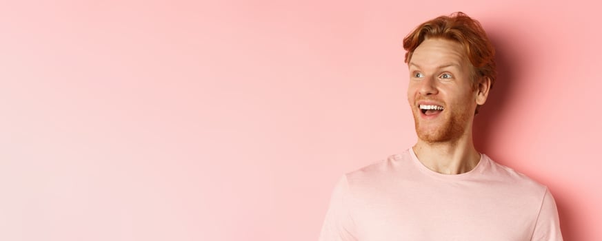Close-up of handsome caucasian guy with red hair and beard, looking left with happy smile, checking out promo offer, standing over pink background photo