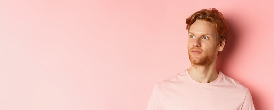 Handsome european male model with red hair and beard, turn head and looking pleased at copy space on left side, standing over pink background photo