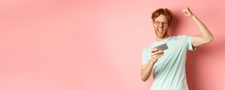 Happy redhead man winning in mobile game, raising hand up and shouting yes with joy, celebrating victory, looking at smartphone, standing over pink background photo
