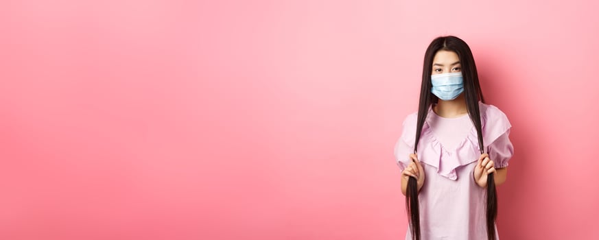 Healthy people and covid-19 pandemic concept. Sassy asian woman in medical mask playing with long hair, looking cool at camera, standing against pink background photo