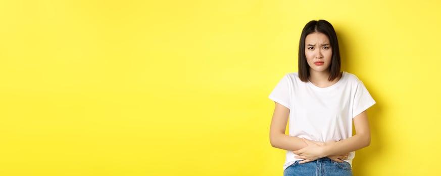 Image of asian woman feeling sick, having painful cramps, holding hands on belly and frowning from pain, discomfort during menstrual period, standing over yellow background photo