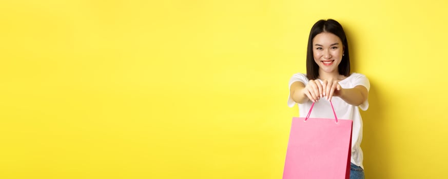 Cute asian girl giving you gift, stretch out hand with pink shopping bag and smiling, congratulating with holiday, standing over yellow background photo