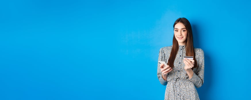 Online shopping. Happy smiling woman buying in app, holding smartphone and plastic credit card, blue background photo