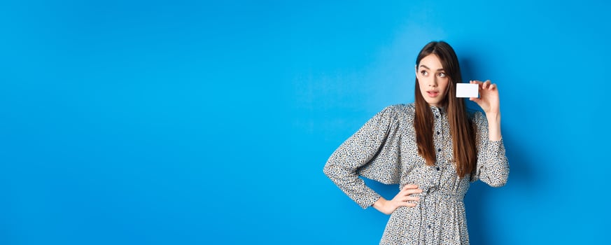 Shopping. Thoughtful female shopper looking left at logo, showing plastic credit card, standing on blue background photo