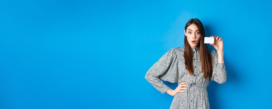 Shopping. Excited girl showing her plastic credit card and look at discounts, say wow amazed, standing on blue background photo