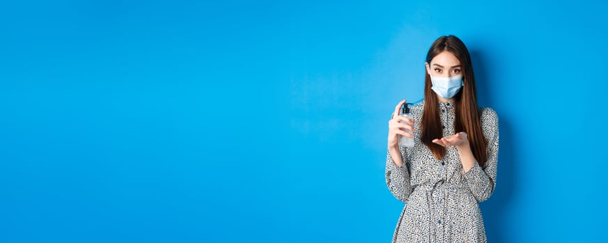 Covid-19, social distancing and healthcare concept. Beautiful girl in medical mask using hand saniziter, holding antiseptic bottle, standing on blue background photo
