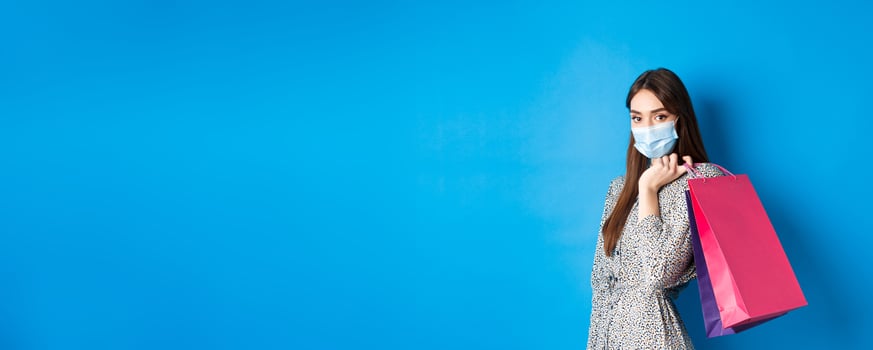 Covid-19, pandemic and lifestyle concept. Elegant woman wear medical mask on shopping, holding bags over shoulder and walking against blue background photo