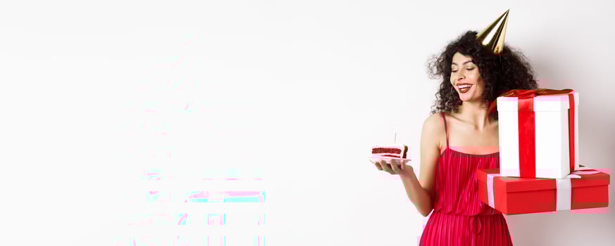 Cheerful smiling woman in red dress and party cone, looking happy at birthday cake, holding gifts, standing on white background photo