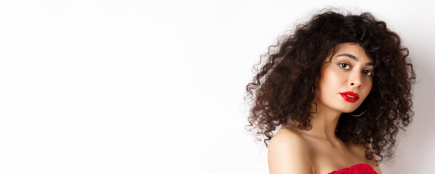 Close-up of beautiful caucasian woman with curly haircut and red lipstick, looking tender at camera, standing over white background photo