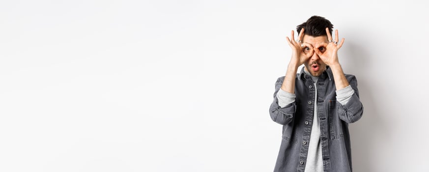 Funny guy look through hand binoculars, checking out awesome promo, say wow and stare at camera excited, standing on white background photo