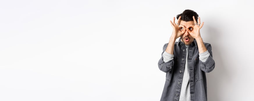 Funny guy look through hand binoculars, checking out awesome promo, say wow and stare at logo on left with excited face, standing on white background photo