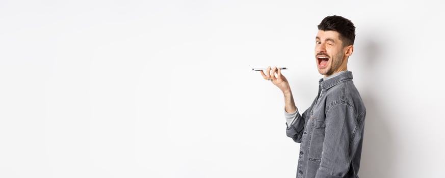 Happy guy winking at looking at camera while using voice translator app or talking on speakerphone, holding phone close to mouth, standing on white background photo