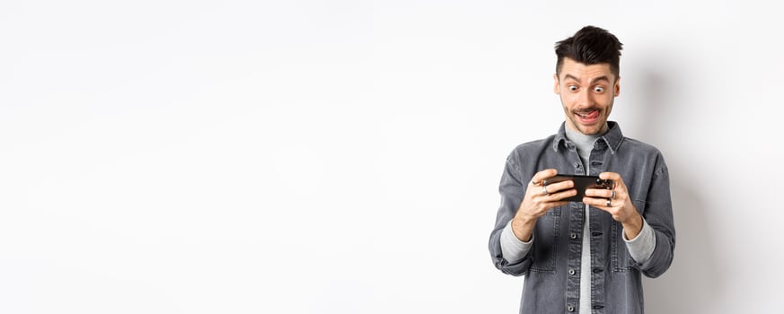 Excited guy playing game on mobile phone with focused face, tap on smartphone screen, holding cellphone horizontally, white background photo