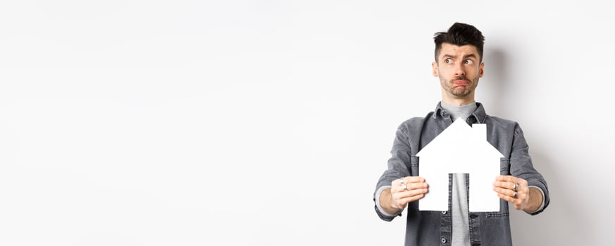 Real estate and insurance concept. Indecisive guy look aside pensive and showing paper house cutout, thinking of buying property, standing against white background photo