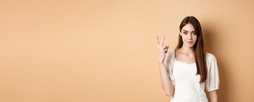 Attractive young woman show fingers number two, smiling and looking confident, standing on beige background photo