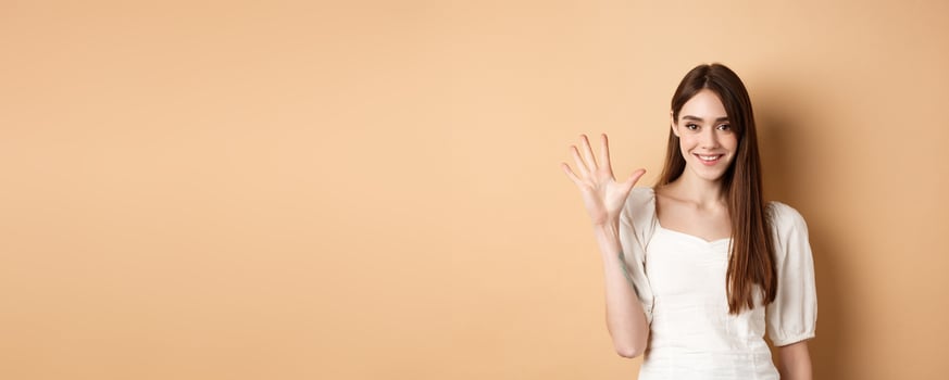 Attractive young woman show fingers number five, smiling and looking confident, standing on beige background photo