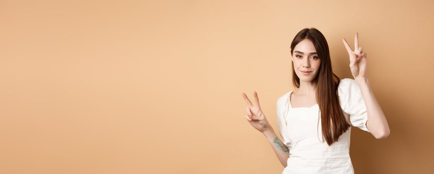 Cheerful attractive girl dancing and having fun, showing v-signs and looking positive at camera, standing on beige background photo