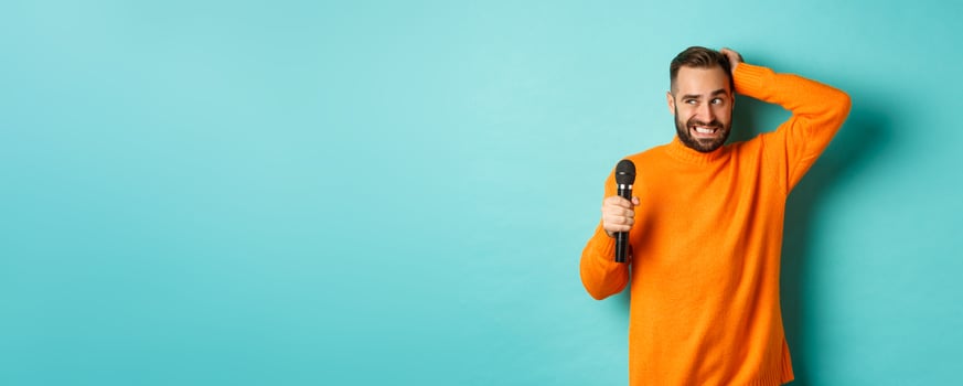Shy and awkward adult man scratching head, holding microphone before singing, standing over blue background photo