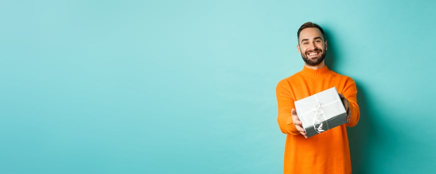 Holidays and celebration concept. Happy man giving you a gift, extending hand with present and smiling, standing over turquoise background photo
