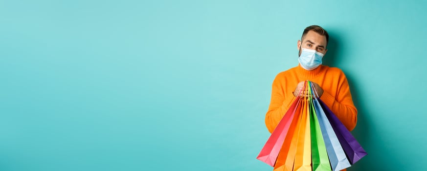 Covid-19, pandemic and lifestyle concept. Displeased and reluctant man in medical mask, holding shopping bags and complaining, standing over turquoise background photo