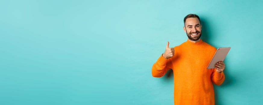 Satisfied adult man smiling, using digital tablet and showing thumb-up, approve and agree, standing against turquoise background photo