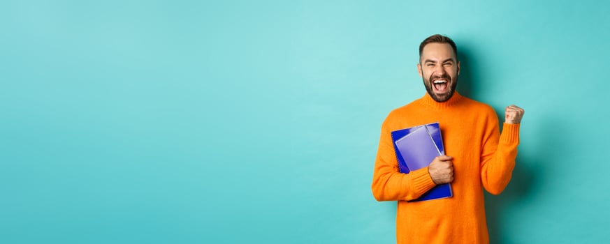 Education. Happy and satisfied man rejoicing, holding notebooks and making fist pump, shouting for joy, standing against turquoise background photo