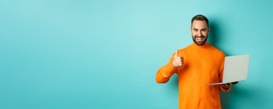 Freelance and technology concept. Lucky man in orange sweater, showing thumb up while working with laptop, standing satisfied over light blue background photo