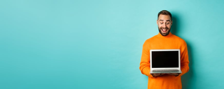 Excited adult male model showing laptop screen, demonstrating online promo, standing happy against light blue background photo