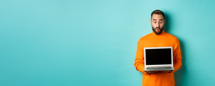 Excited adult male model showing laptop screen, demonstrating online promo, standing happy against light blue background photo