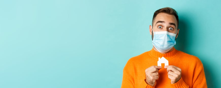 Real estate and coronavirus pandemic concept. Close-up of adult man in medical mask holding small paper house maket and smiling, searching for apartment, light blue background photo