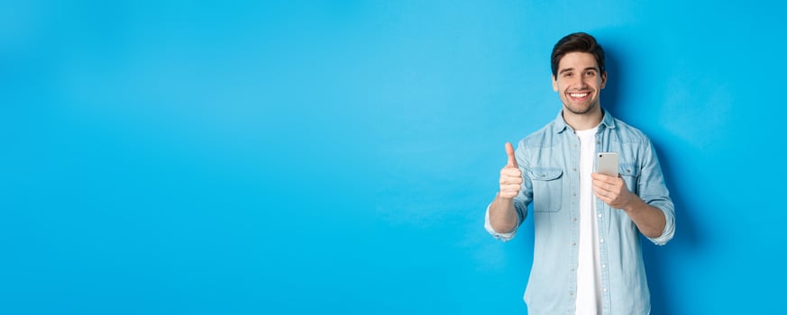 Concept of online shopping, applications and technology. Satisfied man in casual clothes smiling, showing thumbs up after using smartphone app, standing over blue background photo