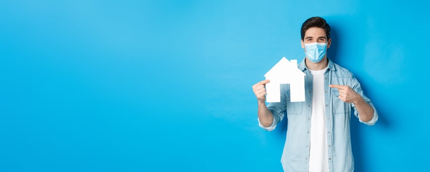 Concept of coronavirus, quarantine and social distancing. Young man searching aparment for rent, business loans, pointing at house model, wearing medical mask, blue background photo