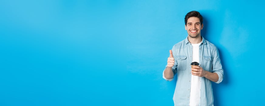 Handsome young man showing thumb-up and drinking coffee, recommending cafe takeaway, standing over blue background photo