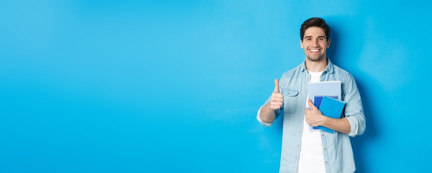 Young man student with notebooks, showing thumb up in approval, smiling satisfied, blue studio background photo