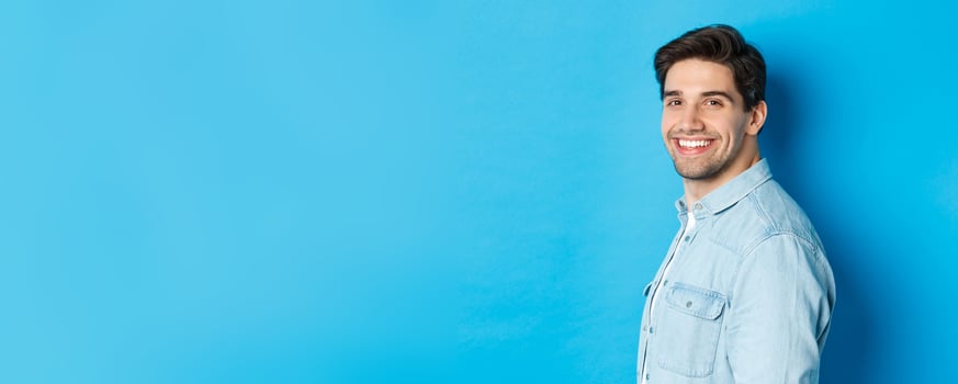 Handsome and confident guy turn head at camera, smiling happy, standing over blue background photo