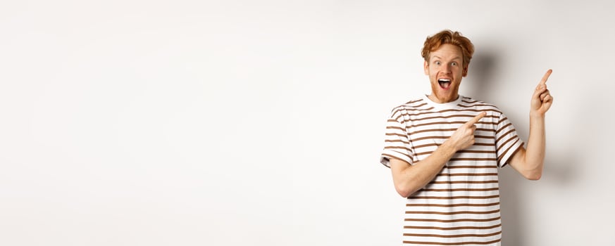 Excited redhead guy in striped t-shirt checking out special deal, pointing fingers right and smiling at camera, white background photo