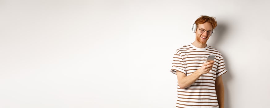 Technology concept. Young redhead hipster in glasses, listening music in headphoes, reading message on smartphone, white background photo