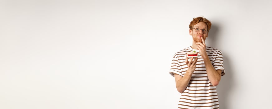 Holidays and celebration concept. Happy young man with red hair and glasses having birthday, holding cake with candle and thinking of b-day wish, standing over white background photo
