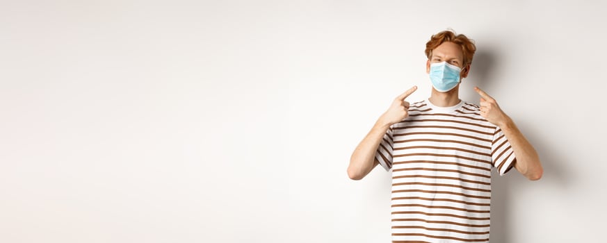 Covid, virus and social distancing concept. Smiling redhead guy pointing fingers at face mask, standing over white background photo