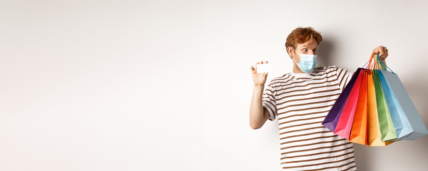 Covid, quarantine and lifestyle concept. Amazed redhead man looking at shopping bags after buying with plastic credit card of bank, white background photo