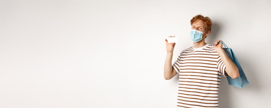 Concept of covid-19 and lifestyle. Happy young shopper in face mask holding shopping bag and showing plastic credit card, buying with discounts, white background photo