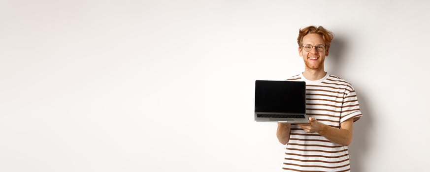 Smiling proud redhead guy showing empty laptop screen. Handsome young man demonstrating logo on computer display, white background photo