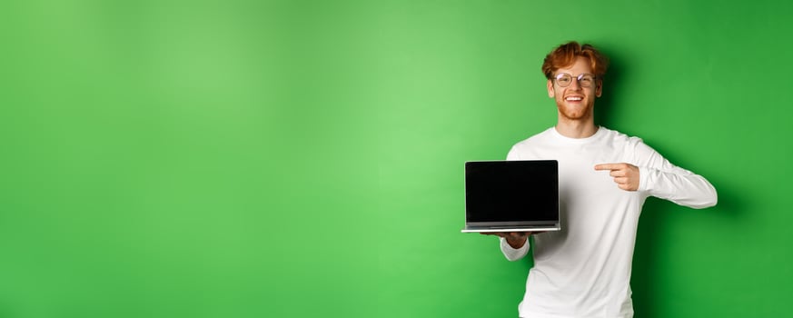 Happy redhead guy in glasses and white long-sleeve t-shirt, pointing finger at blank laptop screen and smiling, standing over green background photo