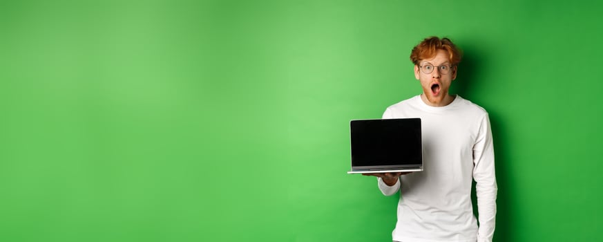 Impressed redhead guy in glasses drop jaw after seeing online promo, showing laptop screen and staring at camera with disbelief, standing over green background photo