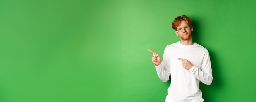 Doubtful redhead guy in glasses pointing fingers left, looking hesitant and unsure at camera, standing over green background photo