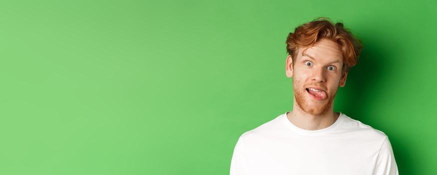 Emotions and fashion concept. Close up of funny redhead man showing silly faces, sticking tongue and staring at camera, standing over green background photo