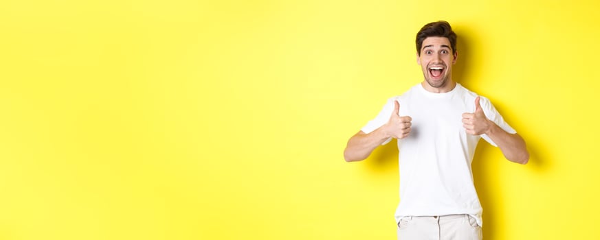 Excited handsome man showing thumbs up, approve and saying yes, standing over yellow background photo