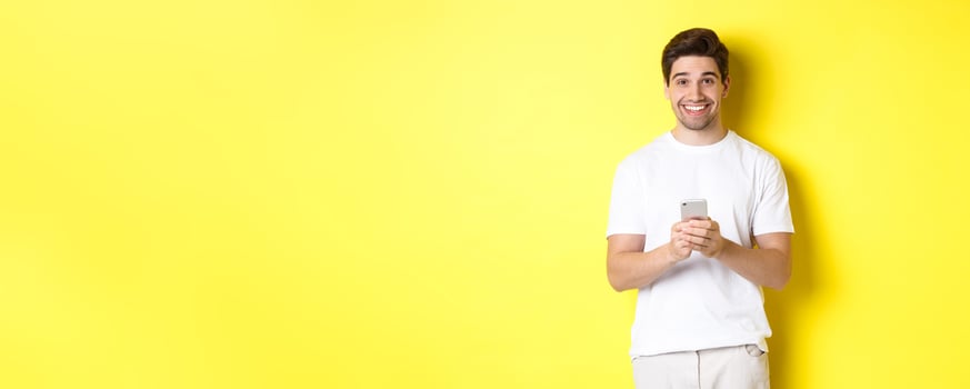 Man smiling and looking happy after reading promo offer on smartphone, standing against yellow background in white t-shirt photo