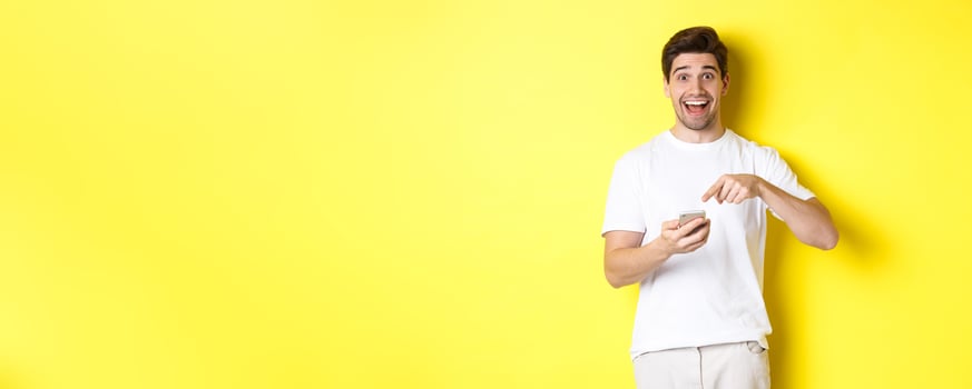 Happy man pointing at smartphone to show promo, check out internet offer, standing over yellow background photo
