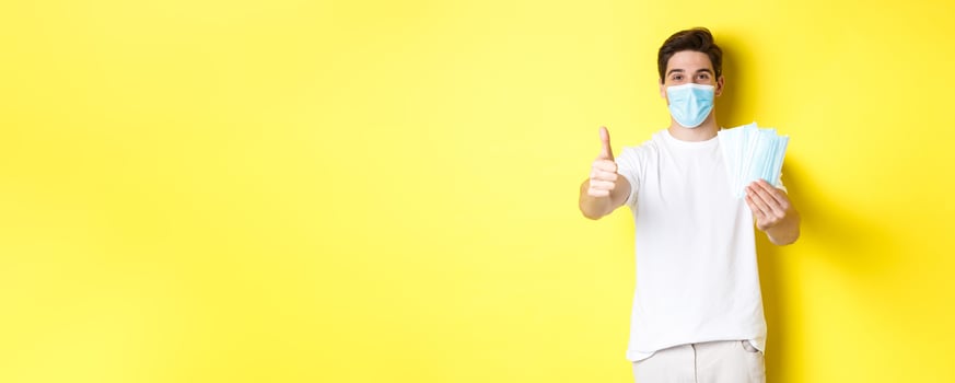 Concept of covid-19, quarantine and preventive measures. Satisfied man showing thumb up and giving medical masks, standing over yellow background photo